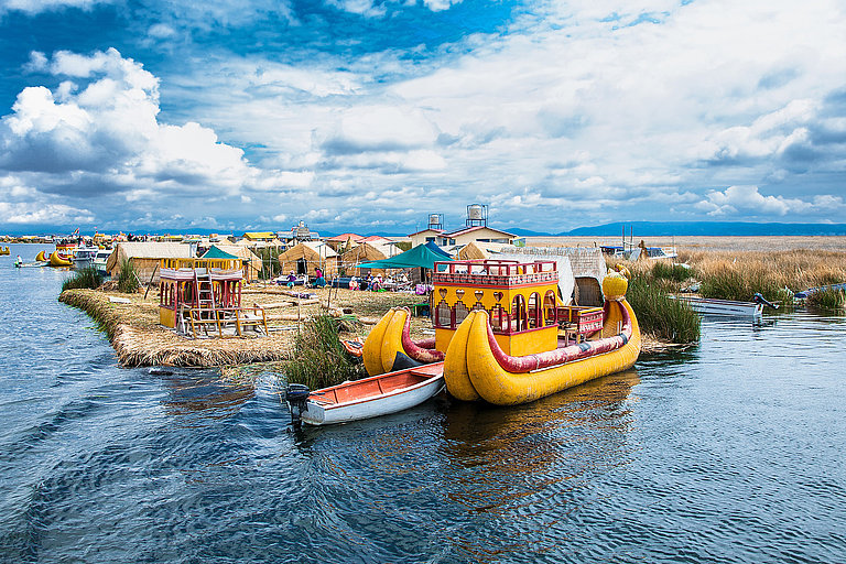 Buntes Holzboot schwimmt auf ruhigem See mit grüner Uferlandschaft und blauem Himmel.