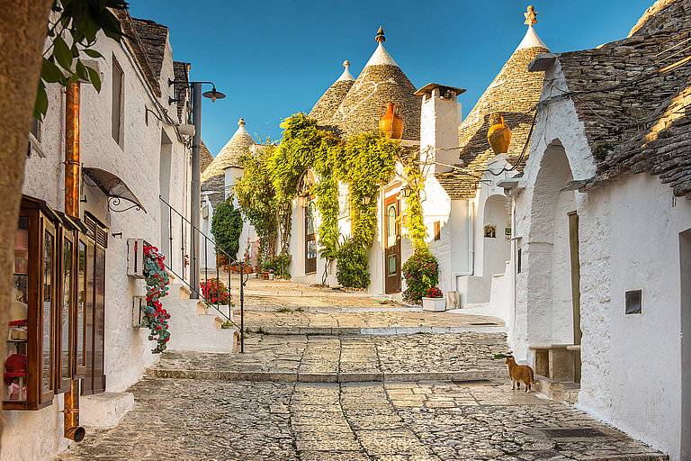 Alberobello Trulli Church, Apulia, Puglia, Italy Kopfsteinpflastergasse mit weißen Häusern und Kegeldächern im warmen Sonnenlicht.