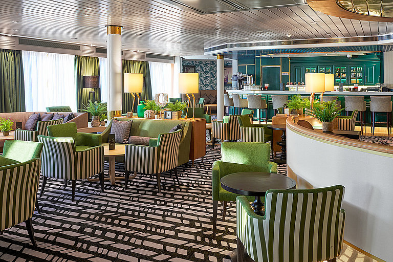 Moderne Schiffsbar mit grün-gestreiften Sitzen, runden Tischen und heller Beleuchtung.