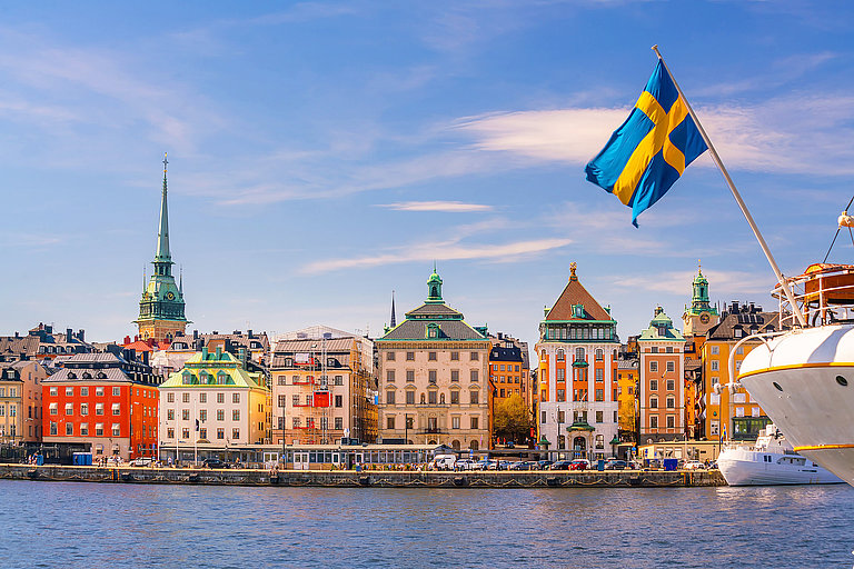 Bunte Altstadt am Wasser mit schwedischen Flaggen und historischen Türmen im Abendlicht.