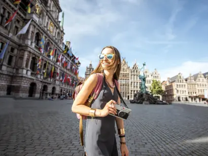 Frau mit Kamera auf historischem Platz mit prachtvollen Gebäuden unter blauem Himmel.
