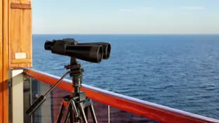 Pinnacle Suite Cat. PS - Binoculars Fernglas an Deck neben der Reling eines Schiffes mit Blick auf das weite blaue Meer.
