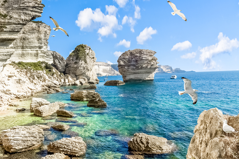 Felsige Küste mit türkisblauem Meer, Booten und markanten Felsen unter Wolken.