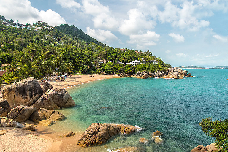 Tropische Küste mit Felsen, Palmenstrand und türkisblauem Meer unter hellem Himmel.