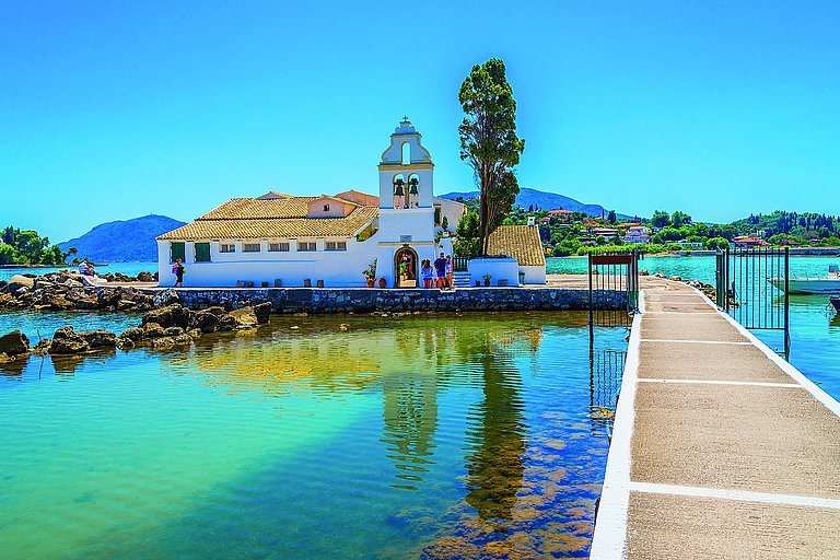 Kleine weiße Kirche auf einer Insel, umgeben von türkisblauem Wasser und Uferweg.