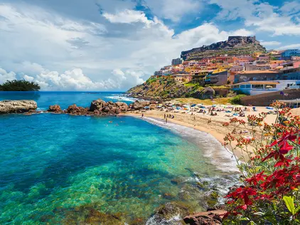 Medieval town of Castelsardo, Province of Sassari, Sardinia, Ita Kiesstrand mit türkisblauem Meer, Blüten und Küstenort vor felsigem Hügel.