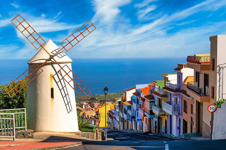 Weiße Windmühle über bunten Häusern mit Meerblick und blauem Himmel.