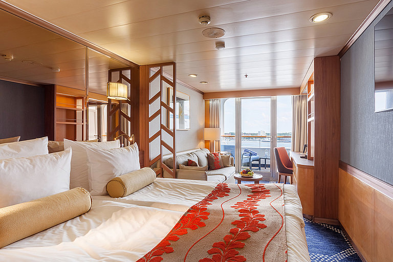 Blick auf helle Schiffskabine auf einem Kreuzfahrtschiff mit rot-beigen Akzenten und Terrasse.