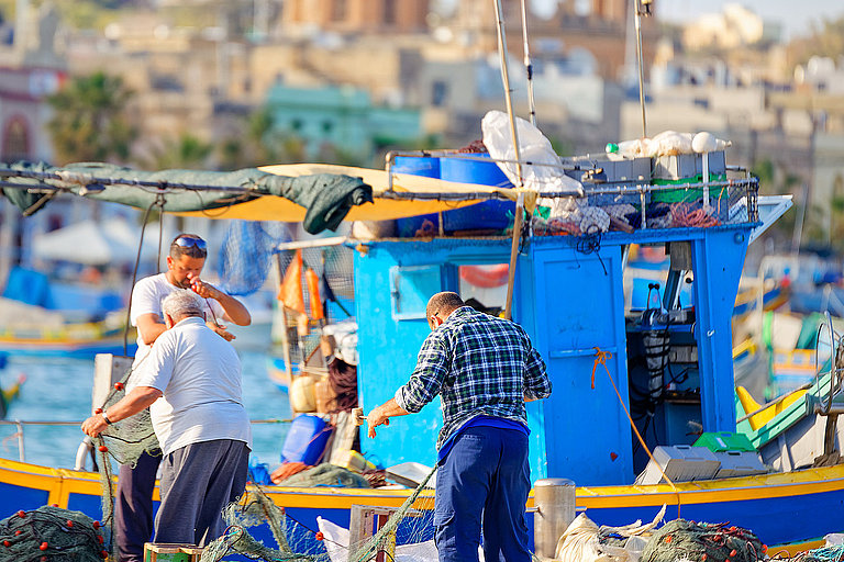 Fishermen from Marsaxlokk, Malta, working their nets Fischerinnen am Hafen mit bunten Booten und historischer Stadtkulisse im Hintergrund.