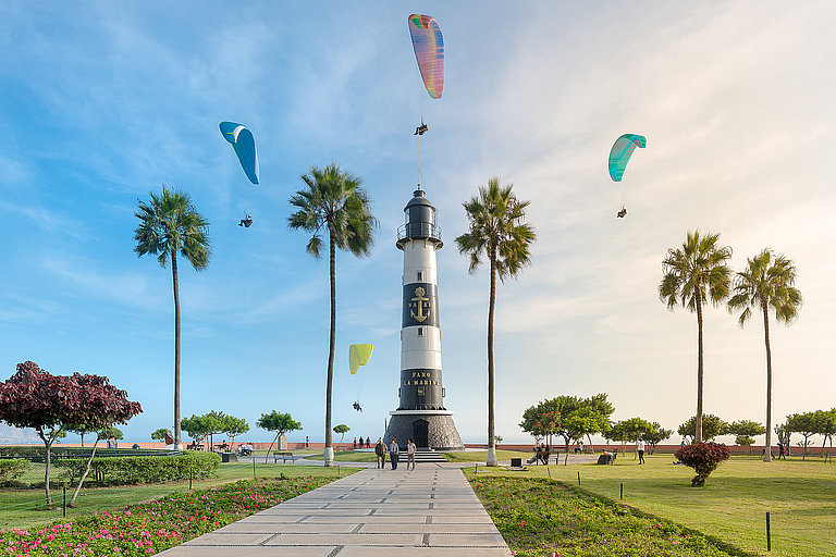 LIMA, PERU: MIRAFLORES LIGHTHOUSE AND PARAGLIDES. Leuchtturm mit Palmen, Blumenbeeten, Tandemspringern und Blick auf das Meer.