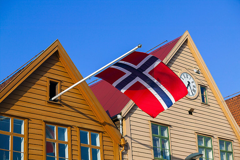 Norwegische Flagge vor Holzhäusern im Bryggen-Viertel unter strahlend blauem Himmel.