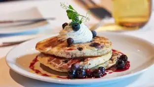 Elegant angerichtetes Dessert mit Pancakes, Creme, Beerensauce und Kresse auf weißem Teller.