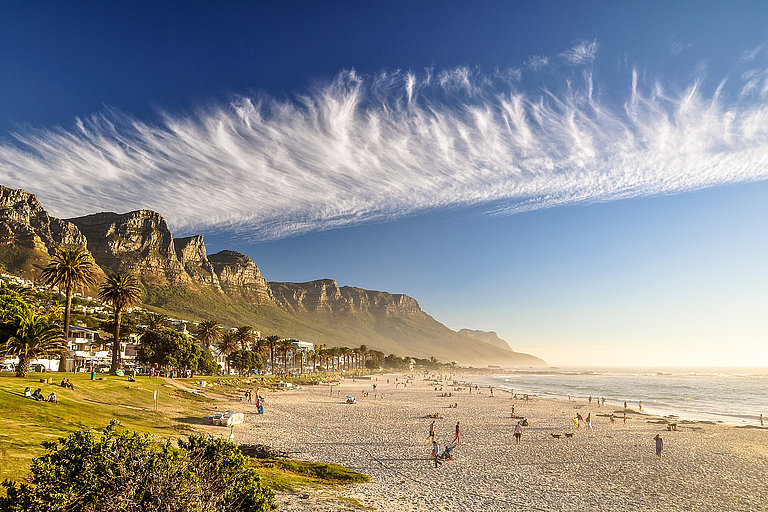 Sandstrand von Kapstadt mit Palmen, Meer und Tafelberg im warmen Abendlicht.