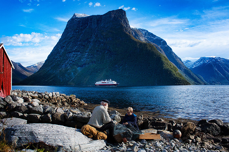 Kreuzfahrtschiff der Hurtigruten fährt auf blauem Meer, vor einer hohen Berglandschaft vorbei.