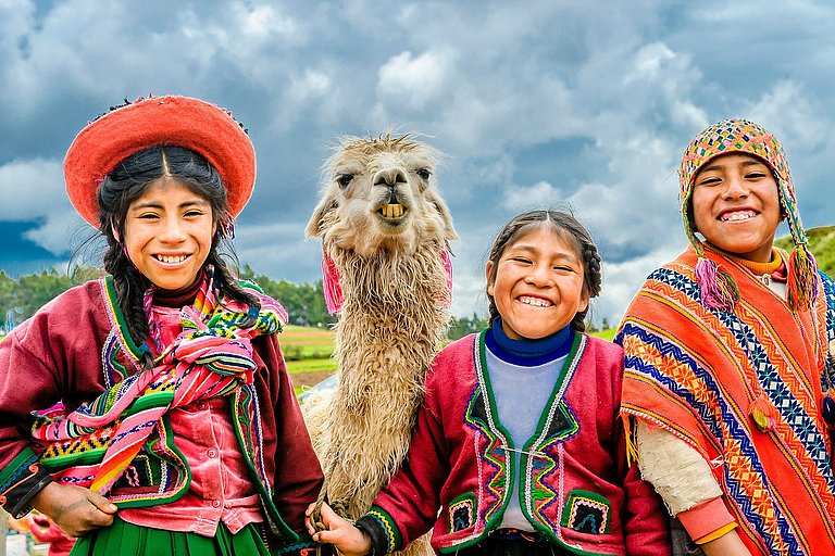 Lächelnde Kinder in bunter Andentracht mit Alpaka vor blauem Himmel in Peru.