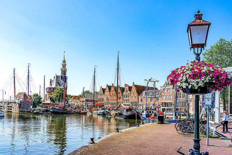 Hafenpromenade mit Segelbooten, Blumen und Altstadt am sonnigen Meer.