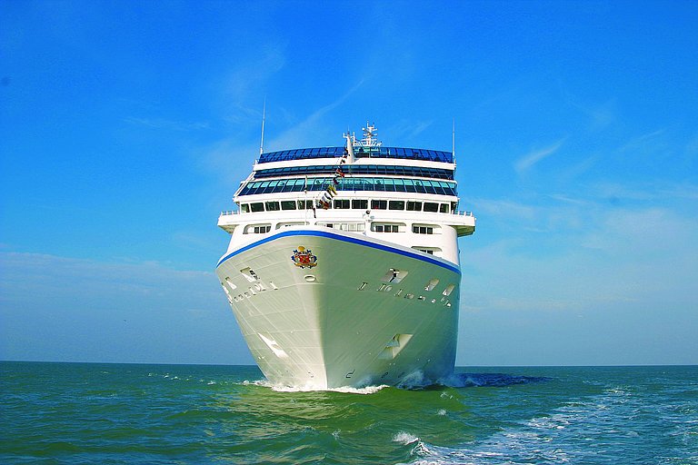 Kreuzfahrtschiff fährt frontal auf blauem Meer unter klarem Himmel mit Bugwelle.