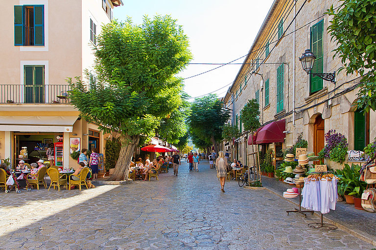 Gepflasterte Gasse mit weißen Häusern, Cafés und grünen Pflanzen im Sonnenlicht.