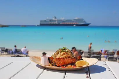 Gericht auf weißem Teller vor türkisblauem Meer und Kreuzfahrtschiff im Hintergrund.