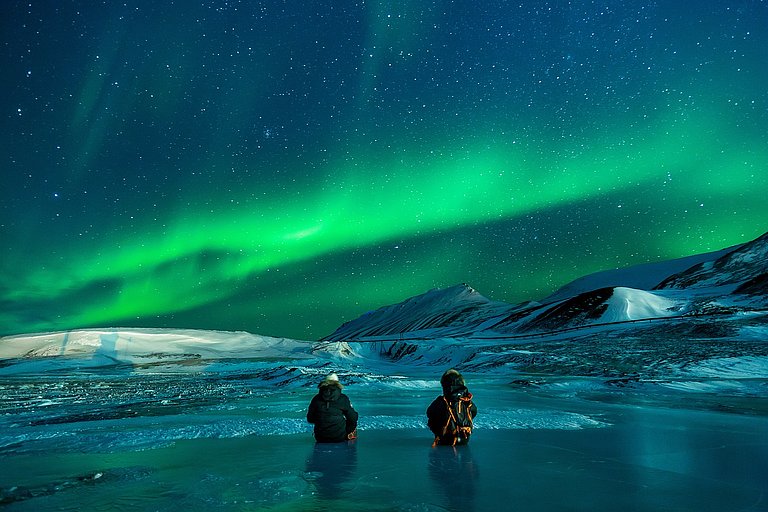 Polarlichter über eisiger Lagune bei Nacht, zwei Personen auf gefrorenem Wasser im Vordergrund.
