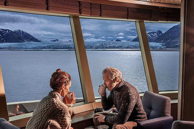 Ehepaar auf einem Kreuzfahrtschiff, sitzt neben einem großen Fenster mit Blick aufs Meer.