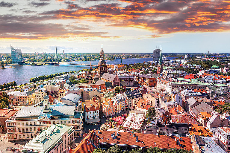 Stadtpanorama mit roten Dächern, Kirchturm und Sonnenuntergang am Horizont.