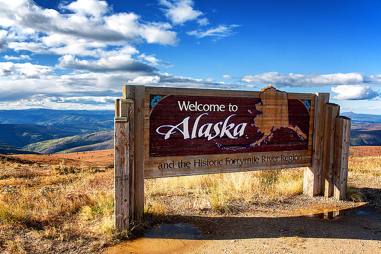 Alaska Welcome Sign Alaska-Willkommensschild vor weiter Küstenlandschaft, Meer und blauem Himmel.