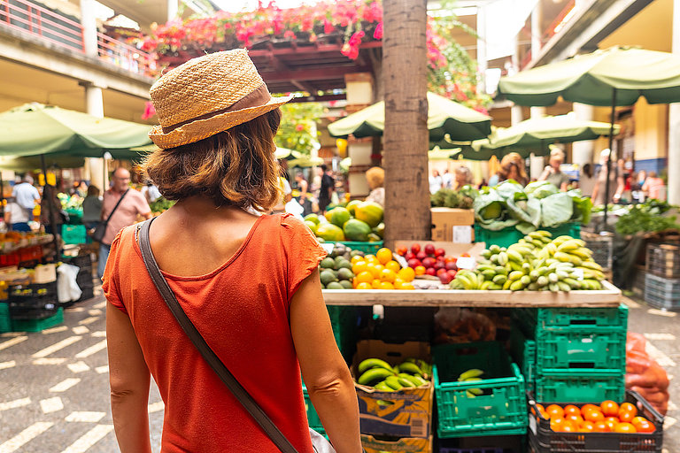 Frau mit Strohhut steht vor einem Markt voller Ständen mit frischem Obst und Gemüse.