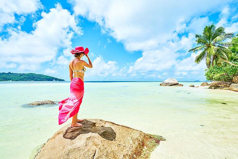 Frau mit Rosa Kleid und Sonnenhut steht auf einem Stein und blickt auf das klare Meerwasser.