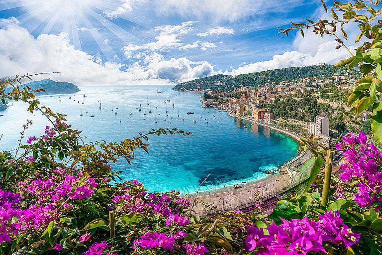 Bucht mit türkisblauem Wasser, blühender Küste und weitem Blick auf Stadt und Meer.