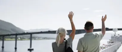 Zwei Erwachsene Personen stehen auf dem Deck eines Kreuzfahrtschiffes und winken.