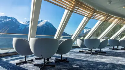 Das Bild zeigt eine Lounge auf einem Kreuzfahrtschiff mit großen Fenstern und Berglandschaft dahinter.