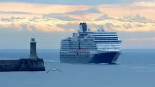 Kreuzfahrtschiff vor Leuchtturm und Hafenmole auf ruhiger See bei bewölktem Abendlicht.