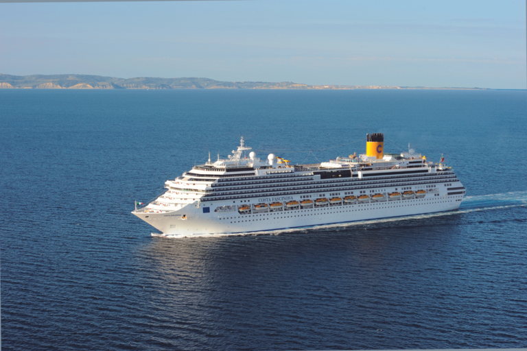 Kreuzfahrtschiff auf blauem Meer, vor wolkenlosen Himmel und der weit entfernten Küste.