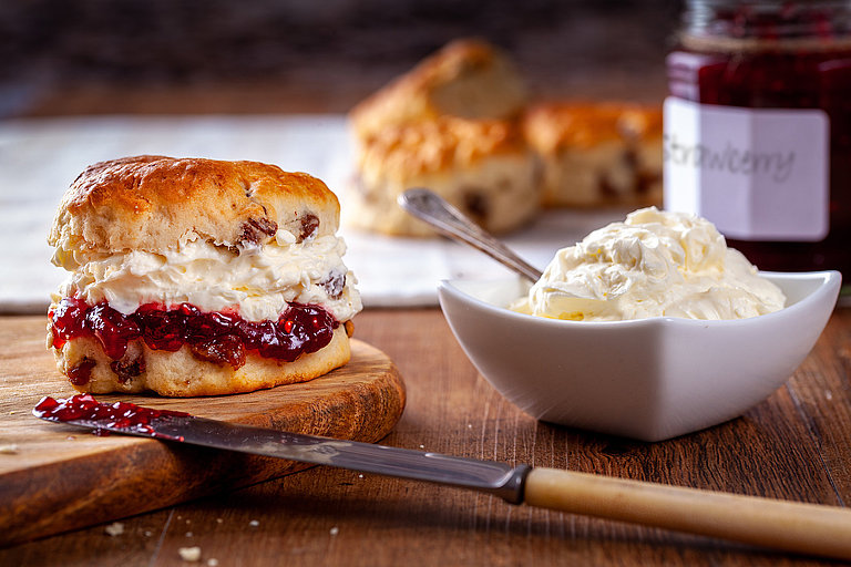 Scone mit Marmelade und Sahne auf Holztisch, daneben Messer und Schälchen.