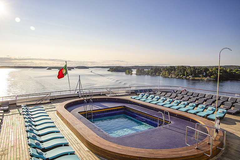 Das Bild zeigt ein Oberdeck mit Pool, Liegen und Blick auf eine grüne Inselgruppe.