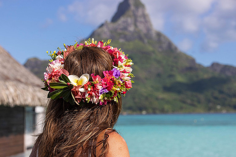 Frau mit Blumenschmuck blickt auf türkisfarbene Lagune und Bergkulisse.