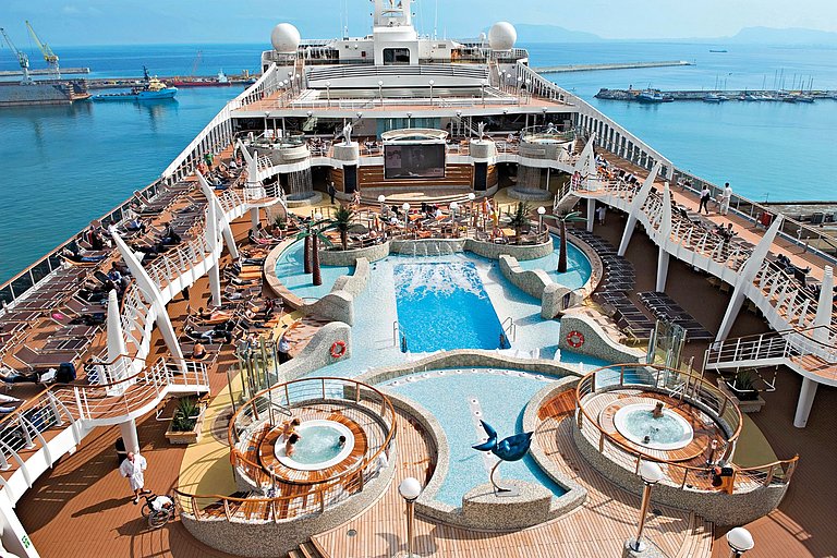 Pooldeck eines Kreuzfahrtschiffs mit Pool, Whirlpools, Plastikpalmen und vielen Sonnenliegen.