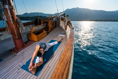 Person sonnt sich auf einem Boot vor glitzerndem Meer und bergiger Küstenlandschaft.