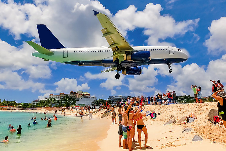 Flugzeug landet spektakulär über einem Strand mit türkisblauem Meer.
