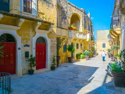 Sonnige mediterrane Altstadtgasse mit Steinhäusern, roten Türen und Pflanzenkübeln.