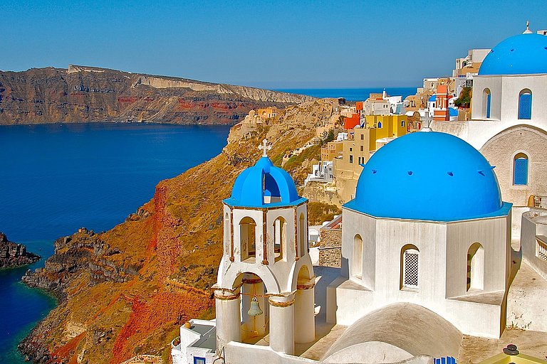 Weiße Häuser mit blauen Kuppeln an der Küste von Santorin über tiefblauem Meer.