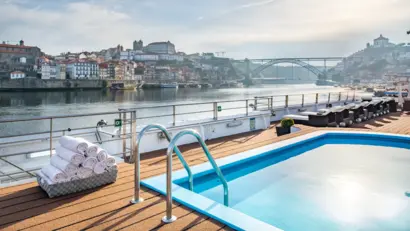 Oberdeck eines Kreuzfahrtschiffs mit Blick auf einen kleinen Pool und eine Stadt mit Brücke.