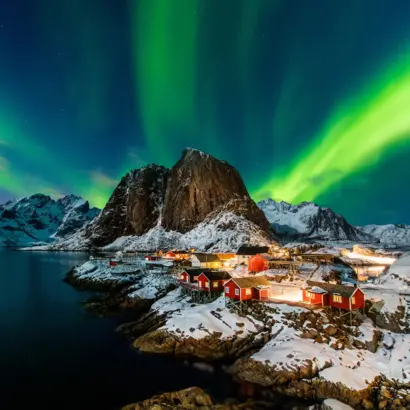 Grüne Nordlichter über verschneitem Lofoten-Dorf, Berg und dunkler Küste.