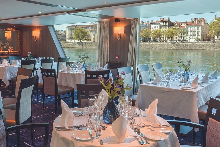 Gedeckte Tische im Restaurant mit großen Fenstern, Tischblumen und Blick auf Flusslandschaft.