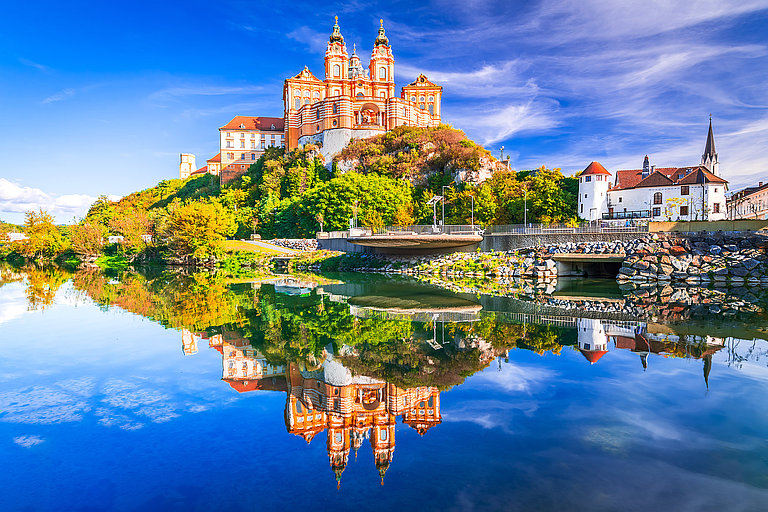 Barockes Stift am Flussufer mit Spiegelungen im Wasser und blauem Himmel.