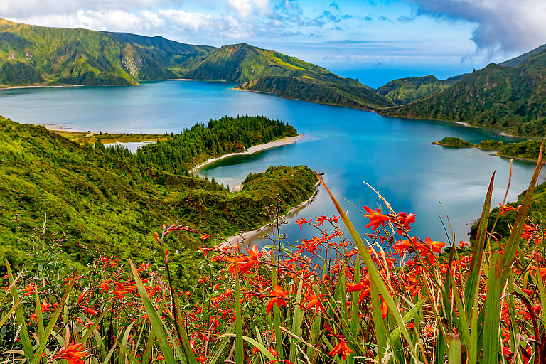 Panoramablick auf die Lagoa do Fogo (Feuersee) auf den Azorhäen, ein tiefblauer Kratersee, umgeben von dicht bewachsenen, grünen Vulkanngen. Im Vordergrund blühen leuchtend orange-rote Montbretien (Crocosmia). 