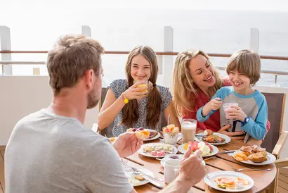 Familie lächelt und sitzt auf einer Terrasse eines Kreuzfahrtschiffes und frühstückt.