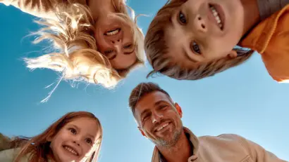 Glückliche Familie blickt lachend in die Kamera, aufgenommen aus der Froschperspektive unter blauem Himmel.