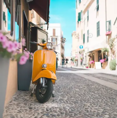 Orangefarbene Vespa in enger Altstadtgasse mit Blumen und Kopsteinpflaster.
